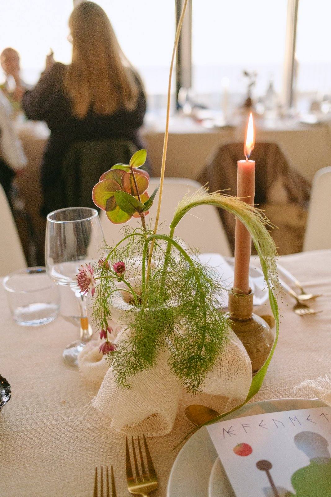 A laid table with a candle, a sculptural arrangement of fennel and a printed menu, guests beyond.