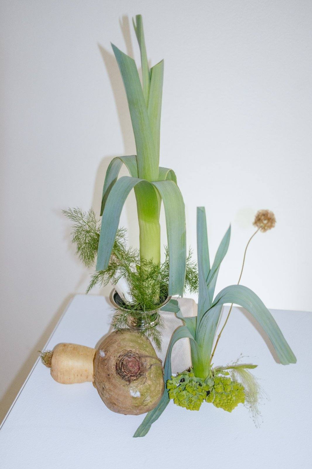 Leeks and a turnip arranged upright in a small vase with yellow flowers.