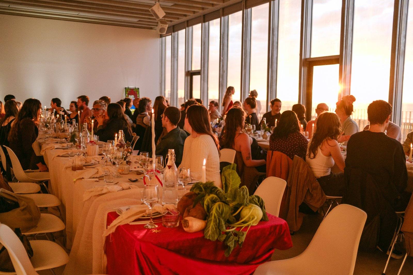 A long banqueting table filled with guests, a red-draped centrepiece of greens in the foreground.