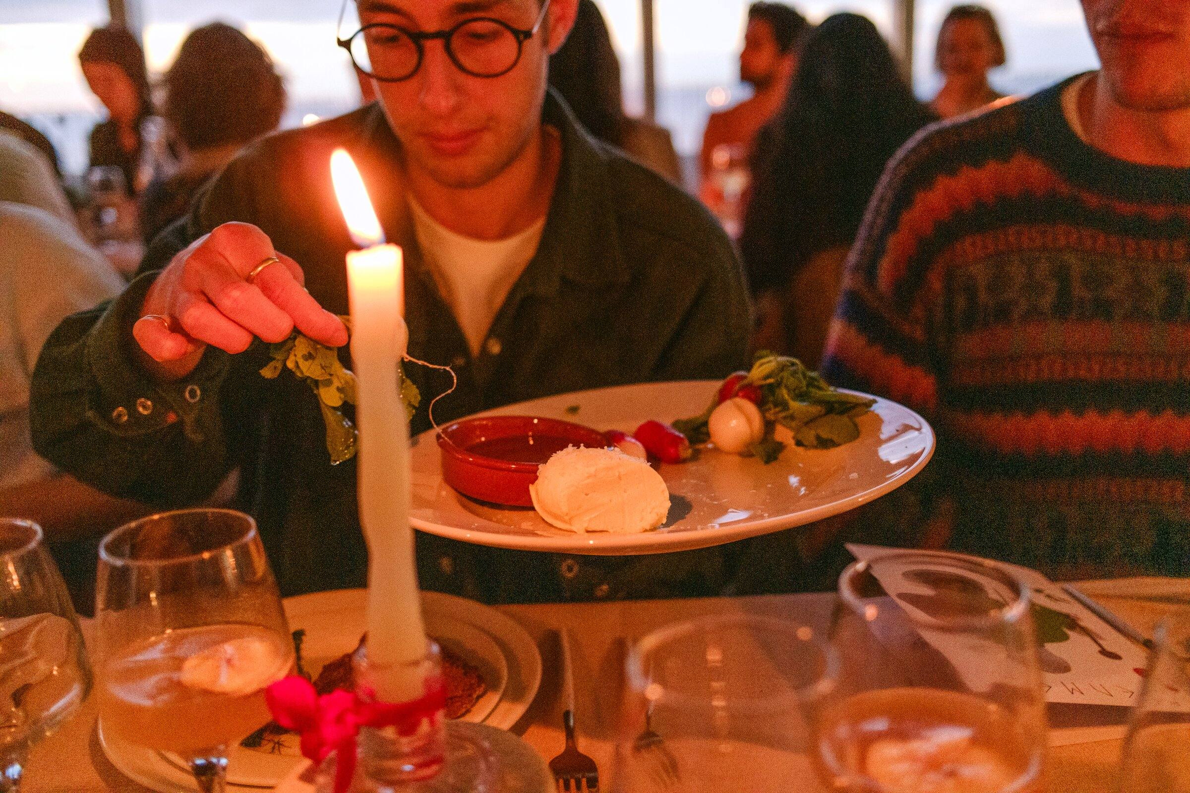 A guest lifts a plate beside a lit candle, surrounded by other diners.