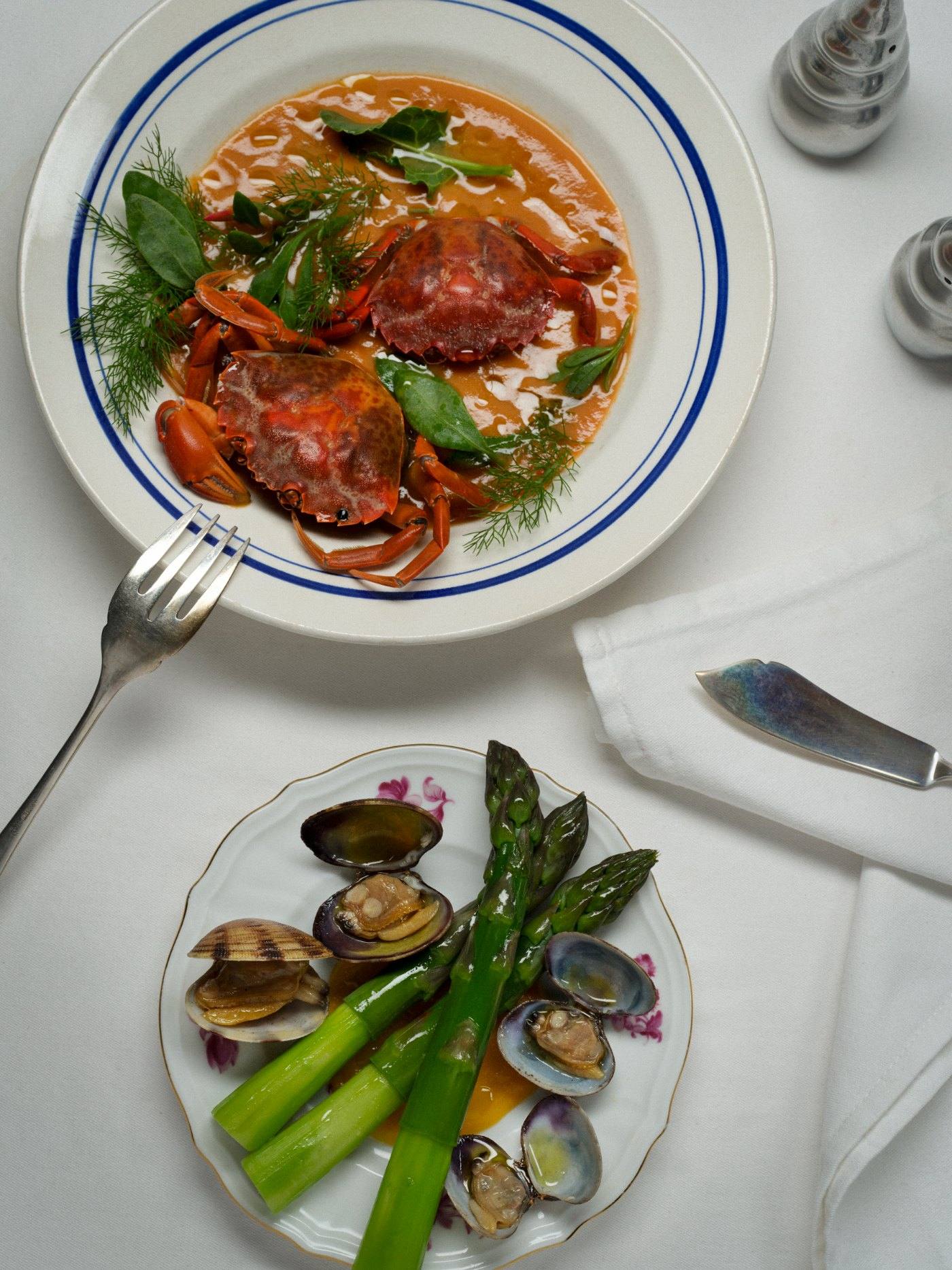 Plates of whole crab in a tomato broth and clams with asparagus on a white tablecloth.